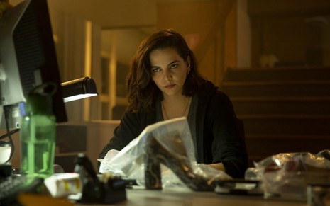 A atriz Tainá Müller, caracterizada como a Verônica da série Bom Dia, Verônica, está sentada em sua mesa de trabalho e olha para a frente com ar de provocação
