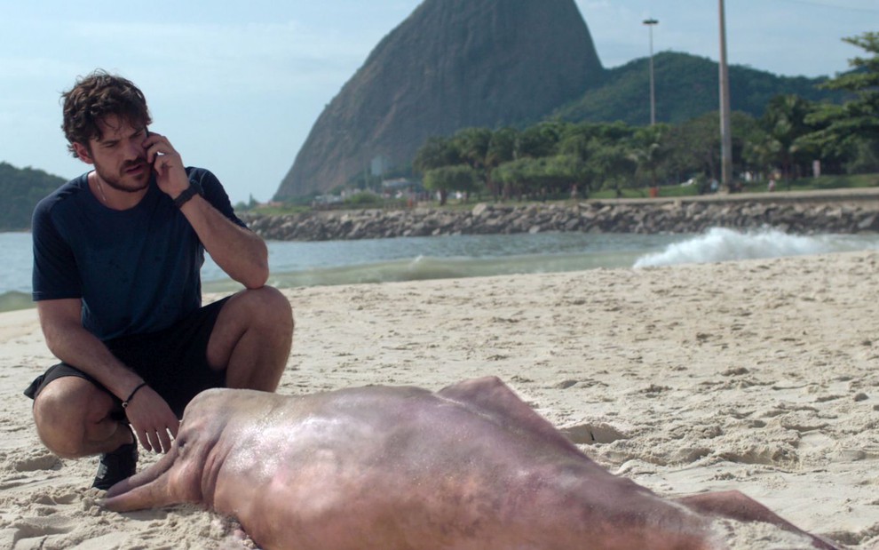 Marco Pigossi fala ao telefone sentado na areia de uma praia em cena da série Cidade Invisível;