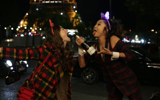 Lily Collins e Ashley Park posam para fotos com bebidas em frente a torre Eiffel na primeira temporada de Emily em Paris