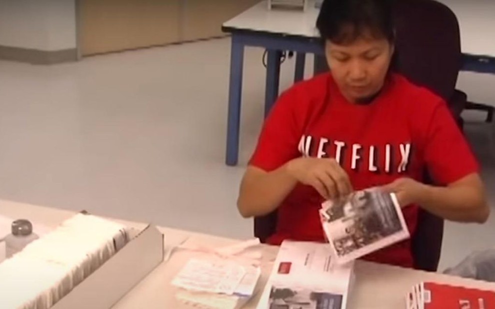 De camisa vermelha com o logotipo da Netflix estampado no peito, funcionária da empresa manuseia um envelope