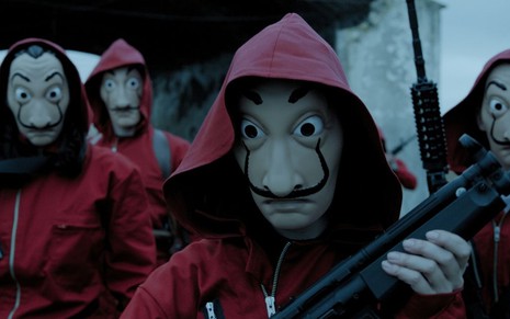 Três mascarados vestem capuzes vermelhos e seguram armas em cena de La Casa de Papel