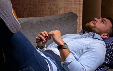 Participante de reality show aparece deitado com as pernas cruzadas em um sofá cinza; ele veste jeans e camisa azul