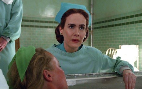 A atriz Sarah Paulson vestida como enfermeira diante de uma paciente em uma banheira como a enfermeira Mildred da série Ratched