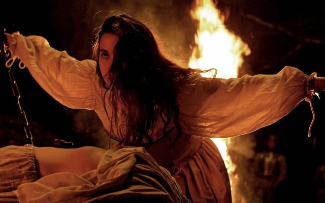 A atriz Amaia Aberasturi com uma veste branca, com os cabelos longos e castanhos na frente da metade esquerda do rosto, ergus os braços diante de uma fogueira como Ana em cena de Silenciadas