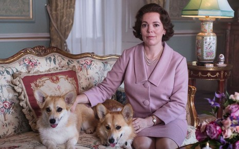 A atriz Olivia Colman sentada com dois cães em um sofá estampado caracterizada como Elizabeth 2ª na terceira temporada de The Crown