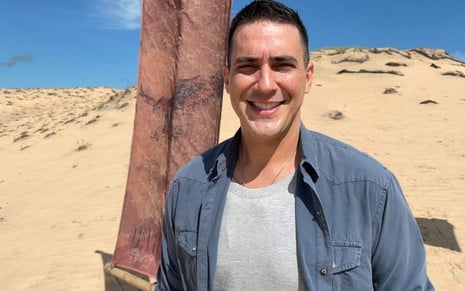 O apresentador Andre Marques sorri; atrás dele, paisagem de dunas e toras de madeira em praia no Ceará