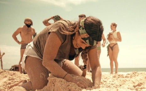 Carol Peixinho cava areia durante prova do No Limite; ao fundo da imagem, participante da equipe Calango conversam