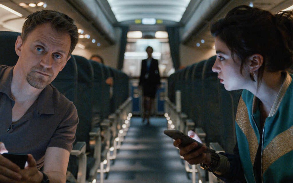 Dentro de um avião, sentados em poltronas à margem do corredor, Vincent Londez e Alba Gaïa Bellugi conversam e mexem no celular em Noite Adentro