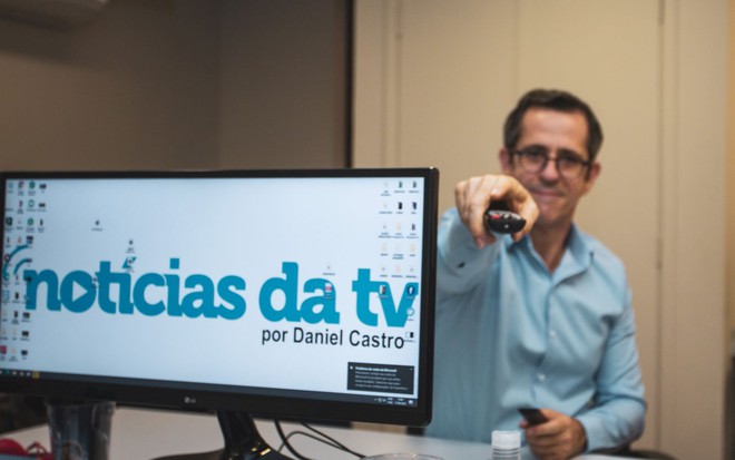 De camisa social azul, Daniel Castro segura um controle remoto na direção da câmera enquanto um monitor de computador exibe a nova marca do Notícias da TV