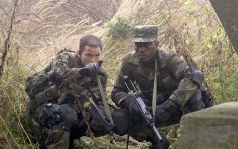 Dois soldados agachados no meio de uma mata, segurando armas; o homem à esquerda esta apontando para alguma coisa fora do quadro