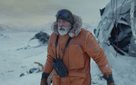 Com um casaco de frio, George Clooney caminha em meio à neve, sua barba está congelada