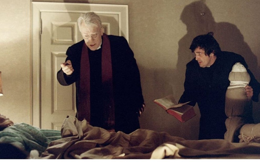 Linda Blair deitada na cama encara os padres de Max von Sydow e Jason Miller em cena de O Exorcista