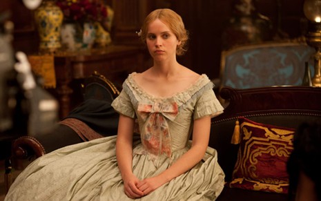 Felicity Jones sentada com um vestido longo de época, com as mãos apoiadas em cima da perna olhando para a frente