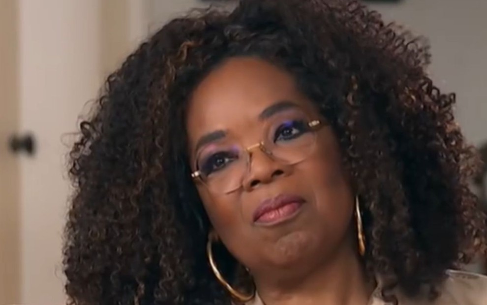 Oprah Winfrey com semblante sério, de óculos de gray, brincos de argola, cabelo solto e blusa perolada em tom marrom claro
