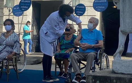 Orlando Drummond de cadeira de rodas, camiseta azul e máscara branca sendo vacinado no braço