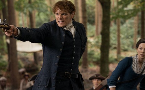 Com uma pistola do século 18 na mão, Sam Heughan protege uma assustada Caitriona Balfe na quarta temporada de Outlander