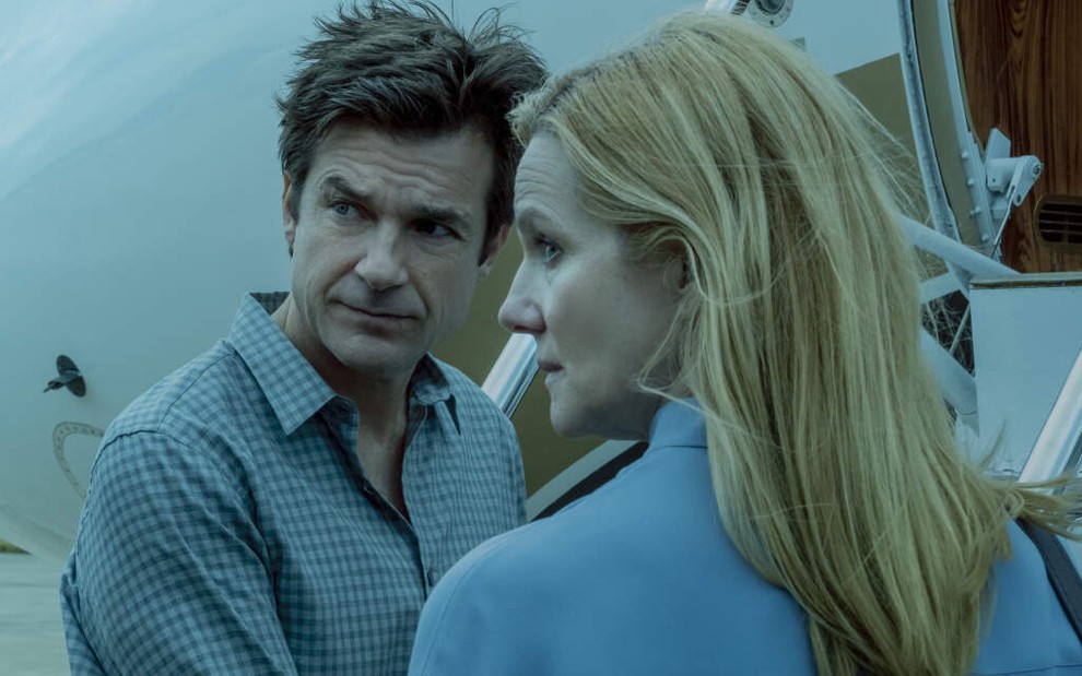 Perto de um avião particular, Jason Bateman e Laura Linney olham para trás em uma imagem com um filtro azul de Ozark, série da Netflix