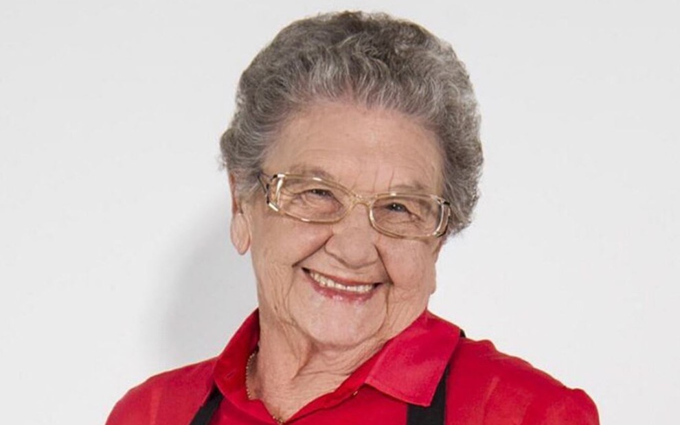 Palmirinha está na frente de uma parede branca, usa blusa vermelha e avental preto; ela está de óculos de grau e sorri para a foto