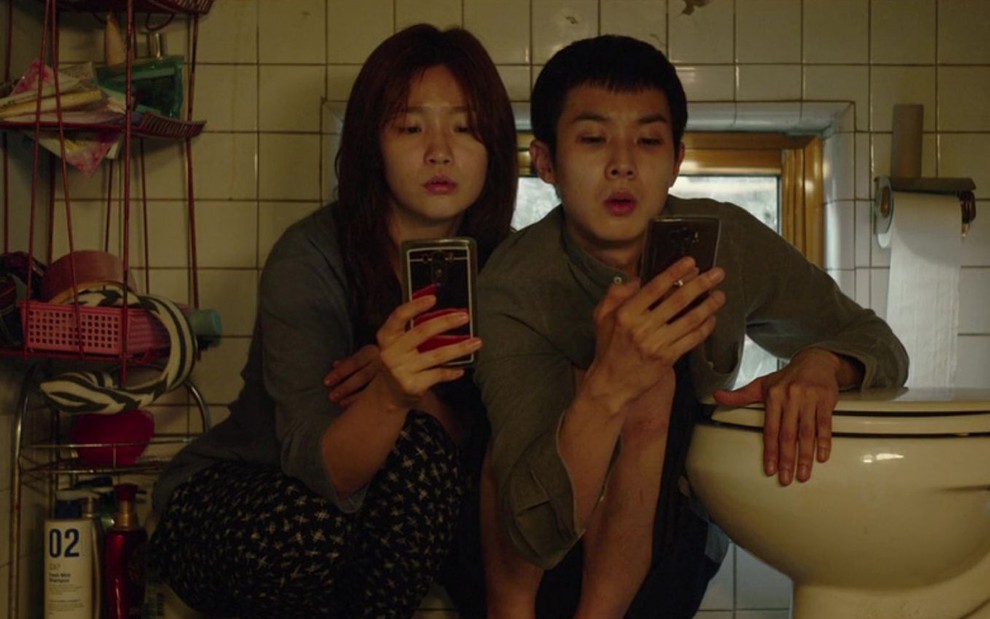 Os atores Park So-dame Choi Woo-sik mexem nos celulares em um banheiro em cena de Parasita (2019)