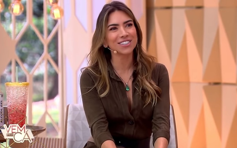 Patricia Abravanel com uma blusa marrom, sorridente, no cenário do Vem Pra Cá