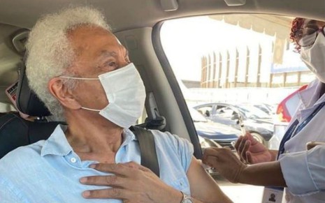 Paulinho da Viola, de máscara em seu carro, recebe injeção da vacina enquanto põe a mão esquerda sobre o peito