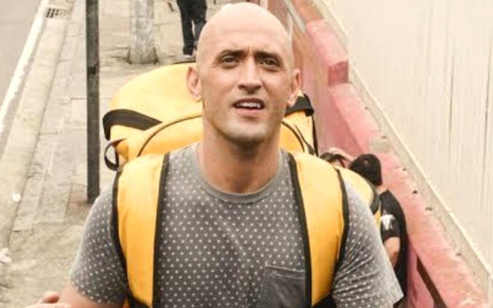 Paulo Gustavo usa uma camiseta cinza e uma mochila amarela em cena como Valdomiro de Vai Que Cola - O Filme