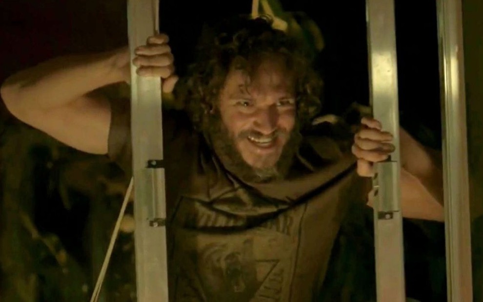 O ator Paulo Rocha abre uma janela; caracterizado como Dino, ele exibe um sorriso macabro em cena de Totalmente Demais