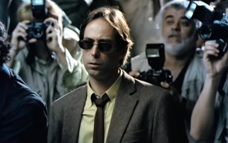 Pedro Cardoso de terno, gravata e óculos escuros, no meio de vários fotógrafos