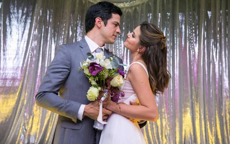 Mateus Solano veste um terno cinza e Camila Queiroz está de vestido branco com um buquê na mão. Eles estão casando-se.
