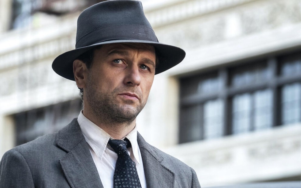 Com um chapéu clássico cinza, que combina com a cor do terno, Matthew Rhys aparece em cena da série Perry Mason