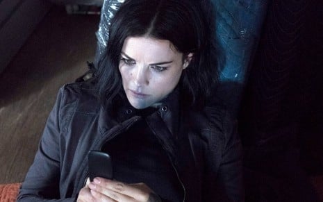 Deitada, Jaimie Alexander olha para um celular antigo e pré-pago na estreia da quinta temporada de Ponto Cego