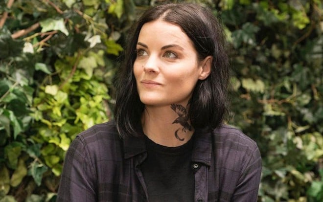 Com umas plantas bem esverdeadas ao fundo, Jaimie Alexander sorri em cena da quinte temporada de Ponto Cego