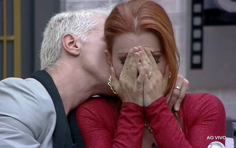 Bruno beija o pescoço de Deborah; ele usa terno cinza e camiseta preta; Deborah está com a cabeça baixa, com as duas mãos no rosto e veste um vestido vermelho