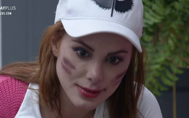 Deborah olha para baixo, ela usa boné branco, blusa rosa com detalhe branco na alça e esta com o cabeço solto; o rosto da participante está pintado com dois riscos marrons