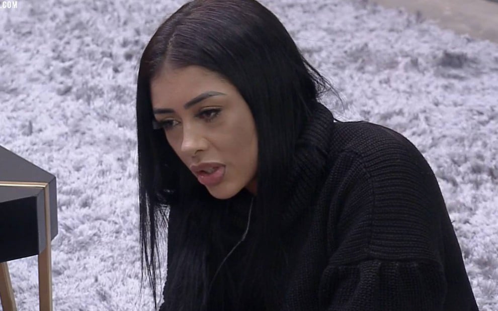 Fernanda Medrado olha para baixo, usa blusa preta e está sentada em um tapete branco