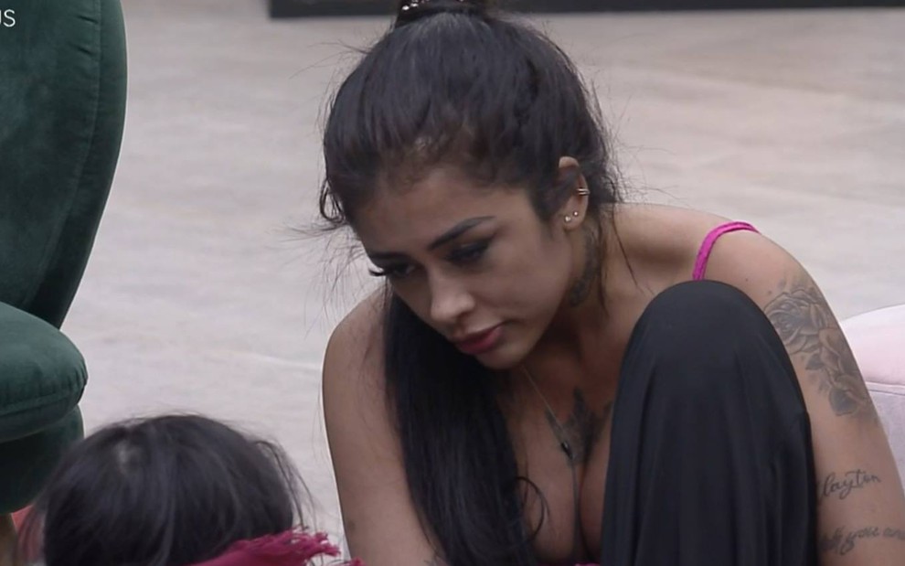 Fernanda olha para baixo, usa biquíni rosa, está com o cabelo presa; a participante está sentada no chão e usa calça preta
