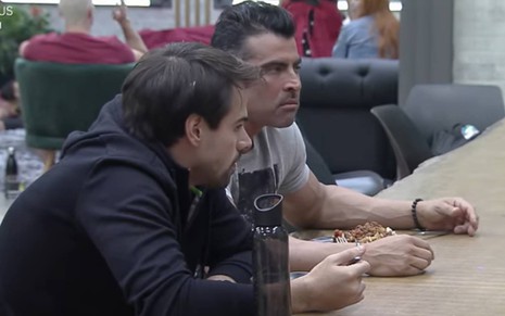 Leandro está sentado ao lado de JP, o marido de Renata usa blusa preta e está com as mãos em cima da mesa; JP está vestindo camiseta cinza e as mãos em cima da mesa
