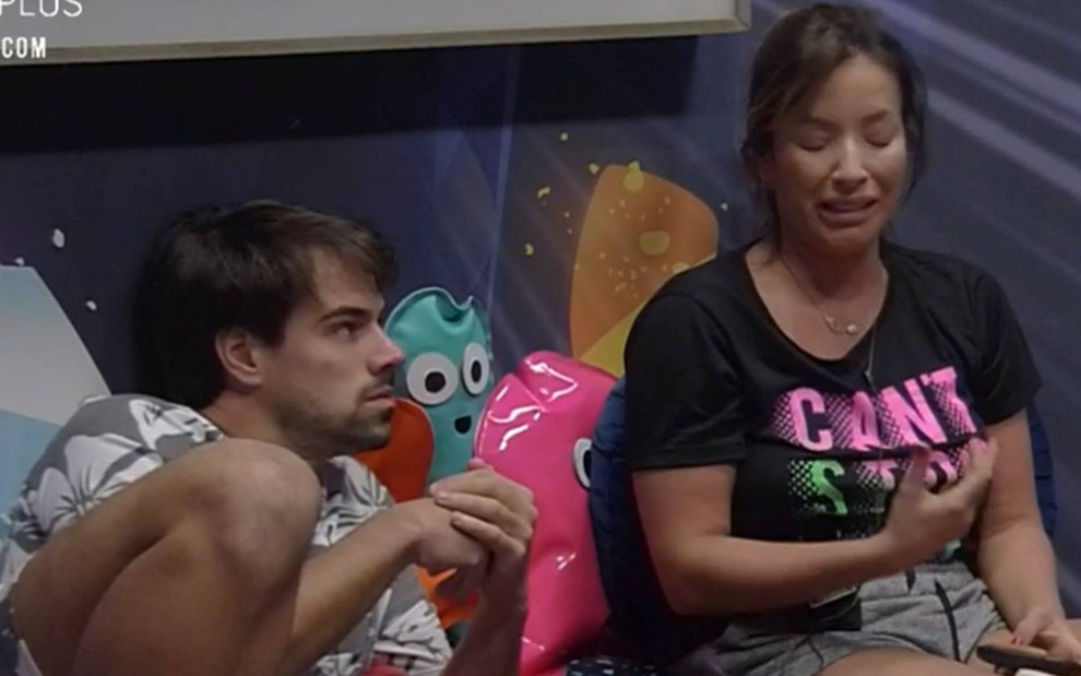 Leandro e Renata estão sentados na cama; ele usa camiseta cinza com flores brancas e olha para a mulher; Renata olha para baixo e chora; ela usa camiseta preta com a frase 'can't stop'