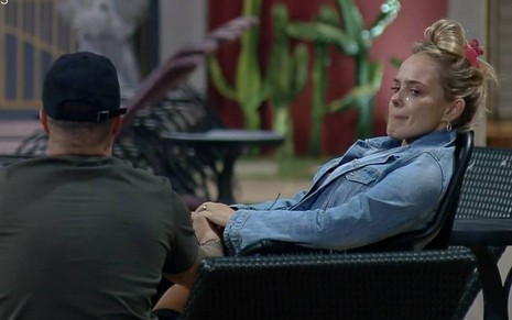 Filipe está de costas, sentado, ele usa camiseta preta e boné preto; Nina usa jaqueta jeans azul e está sentada, ela chora e olha para o companheiro