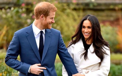 Príncipe Harry de terno azul, olhando sorridente para Meghan Markle, de casaco branco
