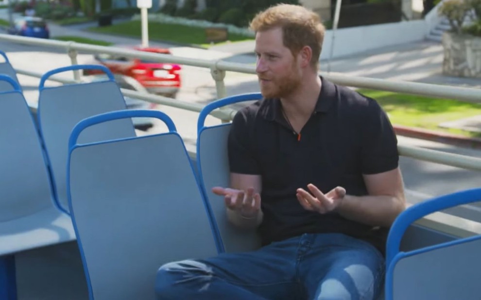 Príncipe Harry gesticula com as mãos sentado no segundo andar de um ônibus aberto com estofado azul nas cadeiras