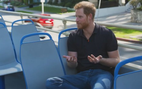 Príncipe Harry gesticula com as mãos sentado no segundo andar de um ônibus aberto com estofado azul nas cadeiras