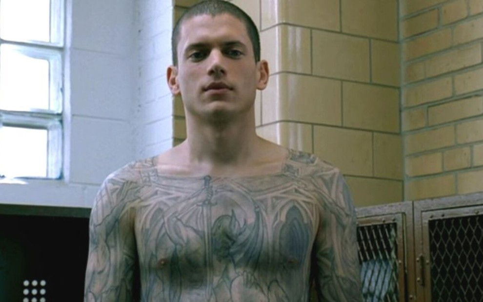 Em um vestiário, Wentworth Miller tira a camisa para mostrar o torso e braços cheios de tatuagens em Prison Break
