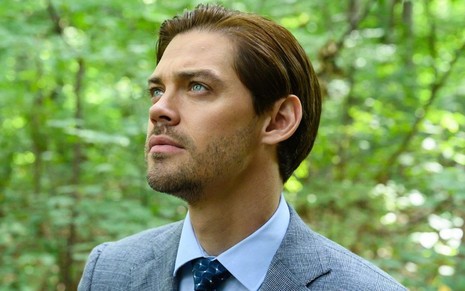 Dentro de uma floresta, Tom Payne aparece veste paletó cinza bem alinhado e esbanja um penteado curto em Prodigal Son