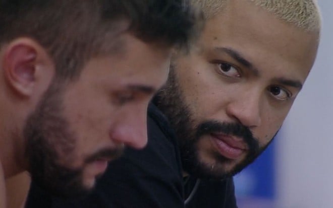 Projota, ao fundo, olha para o rosto de Arthur, cabisbaixo e desfocado, na parte externa do BBB21