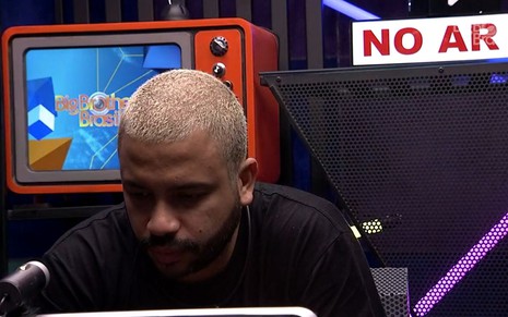 O rapper Projota cabisbaixo sentado no confessionário do BBB21, com uma TV com o logotipo do Big Brother Brasil ao fundo