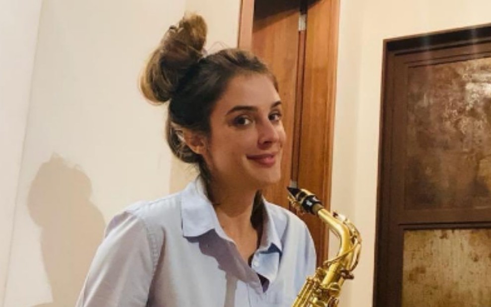 A apresentadora Rafa Brites em sua casa de cabelo preso e segurando um saxofone