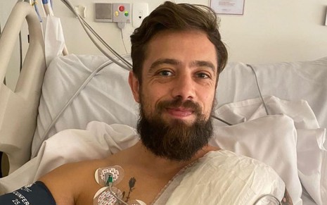 O ator Rafael Cardoso, deitado em uma maca de hospital com curativos ao redor do corpo, logo depois de realizar cirurgia