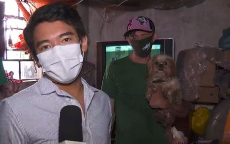 Rafael Ihara de máscara branca, camisa cinza; entrevistado ao fundo de camiseta verde com cachorro no colo