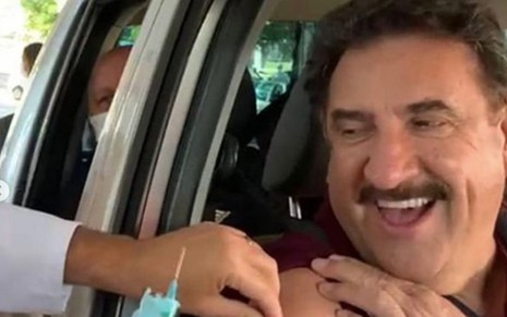 O apresentador Ratinho, sem máscara, dentro de um carro enquanto recebe uma injeção no braço direito